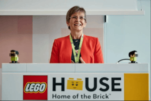 The image shows a person standing behind a podium with a 'LEGO HOUSE - Home of the Brick' sign. The person is wearing a red blazer and a green lanyard.
