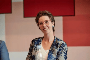 The image shows a woman with short hair, wearing a floral blazer over a light-colored top. She has glasses on and is smiling. The background includes a red wall with two red panels.