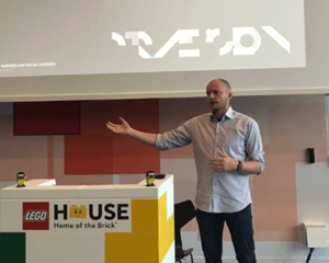 The image shows a person standing in front of a podium with a LEGO sign that says 'Home of the Brick'. Above the podium is a projected image of two white birds flying. The person appears to be giving a presentation or lecture.
