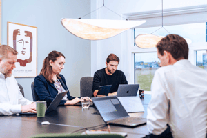 The image shows a professional meeting in progress with four individuals that work at UNIwise around a table. Each person is using a laptop, suggesting a business or collaborative work environment.