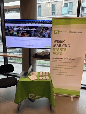 The image shows a booth for UNIwise Marking Studio at an indoor location with a large screen displaying the website and a printed poster beside it listing features of the service.